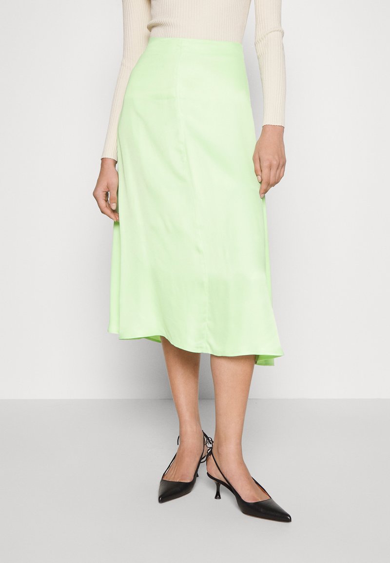 Jupe midi en satin vert avec une texture lisse, forme en A et une légère brillance, associée à des escarpins noirs à bout pointu.