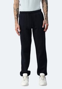 Schwarze Fleece-Jogginghosen mit elastischem Bund und Kordelzug, ausgestattet mit Seitentaschen und schmal zulaufenden Bündchen, kombiniert mit weißen Sneakers.