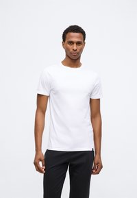 Férfi sima, fehér rövid ujjú pólót és fekete nadrágot visel, miközben egy sima, világosszürke háttér előtt áll.