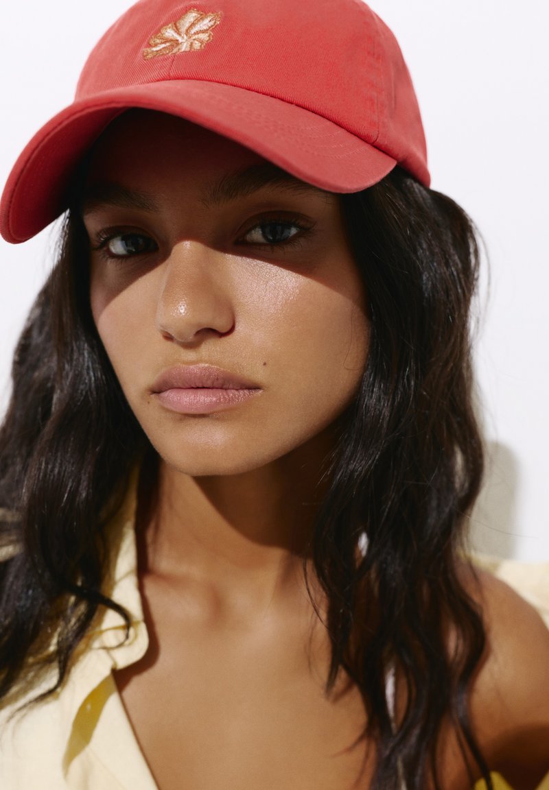 Jeune femme aux longs cheveux foncés portant une casquette rouge avec un emblème de feuille dorée et une chemise jaune clair sur un fond neutre.