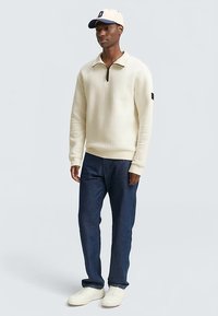 Krémový pletený pulovr s polovičním zipem, žebrovanými manžetami a malým černým detailem na rukávu, doplněný tmavými džíny a bílými teniskami.
