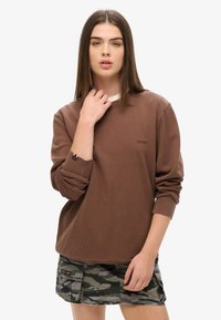 Maglietta marrone a maniche lunghe con scollo rotondo, caratterizzata da un logo sottile e polsini risvoltati. Abbinate a pantaloncini con motivo mimetico.