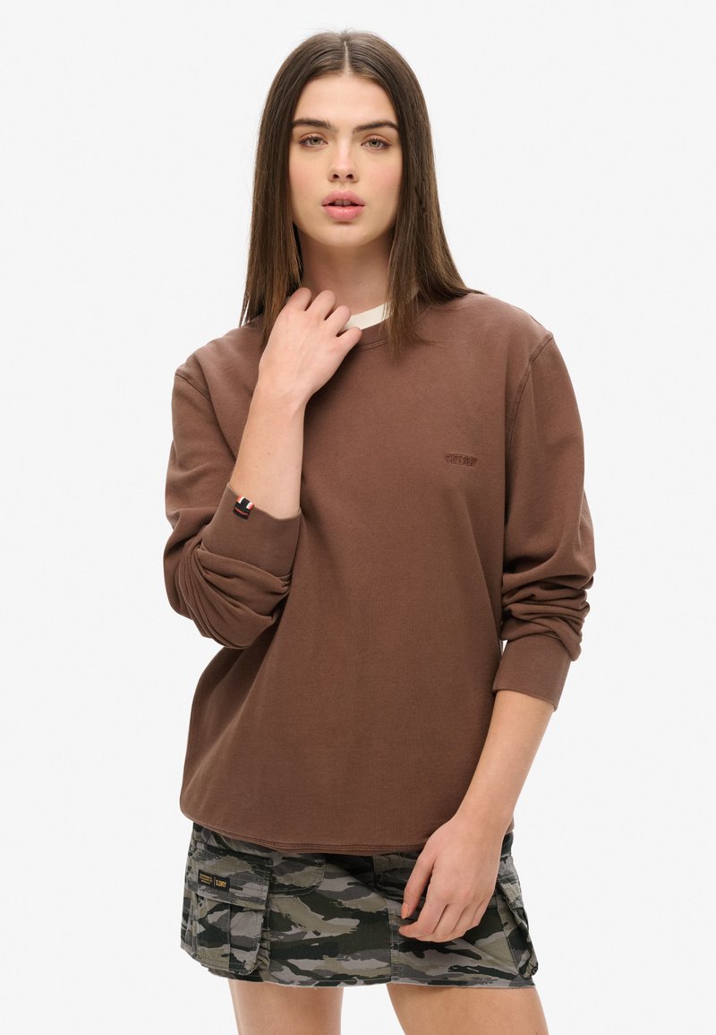 Maglietta marrone a maniche lunghe con scollo rotondo, caratterizzata da un logo sottile e polsini risvoltati. Abbinate a pantaloncini con motivo mimetico.