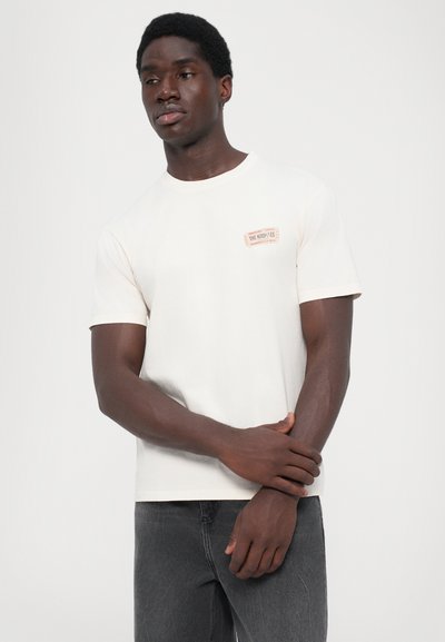 Cream short-sleeve t-shirt with a round neckline and a rectangular label on the upper left chest featuring text. Worn with dark gray jeans.