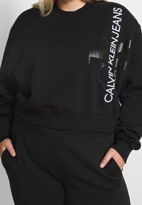 Sudadera negra de tela suave con un diseño recortado. En la parte frontal presenta un logo blanco y acentos gráficos en el lado izquierdo.