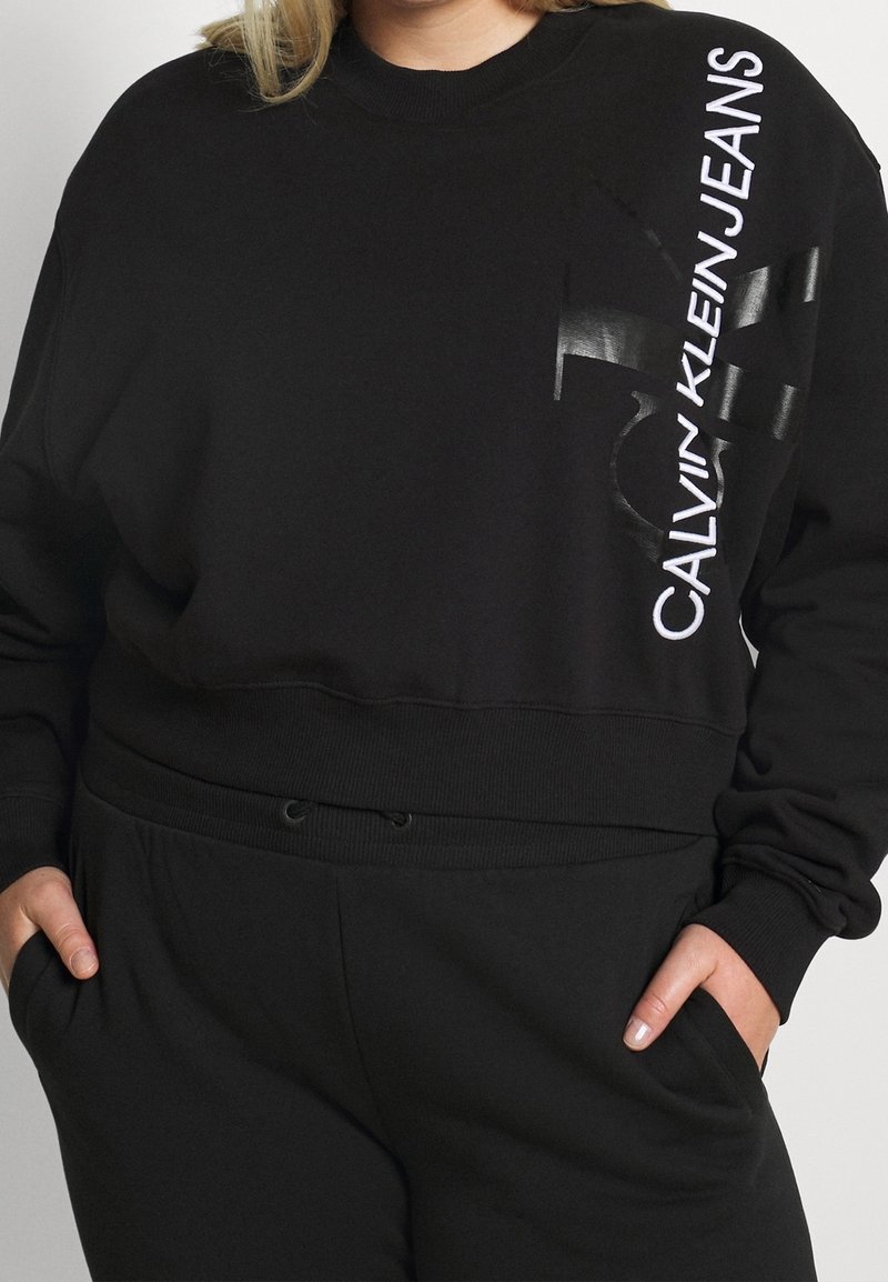 Sudadera negra de tela suave con un diseño recortado. En la parte frontal presenta un logo blanco y acentos gráficos en el lado izquierdo.