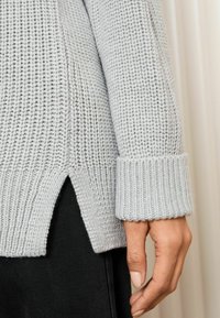 Hellgrauer gestrickter Pullover mit strukturiertem Muster, gerippten Manschetten und seitlichen Schlitzen. Der Stoff scheint weich und gemütlich zu sein.