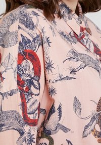 Kortærmet skjorte i lyserød stof med et detaljeret mønster af krybdyr, fugle og planter i blå og røde nuancer. Krave med knaplukning.