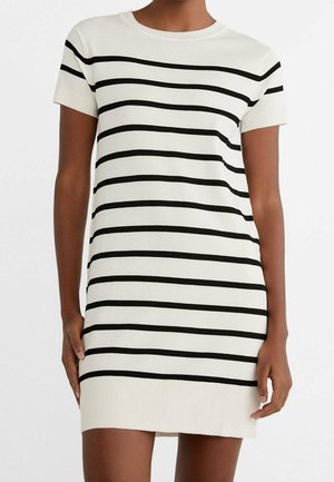 Vrouw draagt een wit gebreide jurk met korte mouwen, voorzien van gelijkmatig verdeelde horizontale zwarte strepen, geribbelde zoom en mouwboorden.