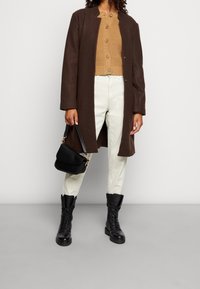 Persona che indossa un cappotto marrone, un maglione beige con bottoni, pantaloni color avorio e stivali neri con lacci, mentre tiene una borsa nera su uno sfondo semplice.