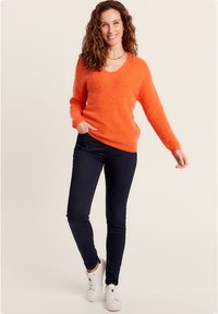 Oranger, flauschiger Pullover mit V-Ausschnitt, langen Ärmeln und gerippten Bündchen, kombiniert mit dunklen Jeans und weißen Turnschuhen. Einfacher und lässiger Stil.