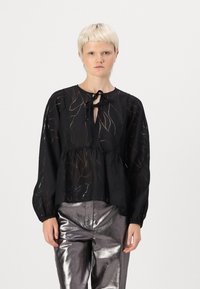 Svart genomskinlig blus med subtilt bladmönster, rund halsringning och bandknäppning; har långa ärmar och en samlad midja. Matchad med glänsande metalliska byxor.