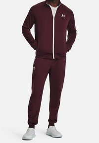 Bordový sportovní komplet vyrobený z měkké tkaniny s vysokým límcem, zapínáním na zip a bílými detaily. V kombinaci se světle šedými teniskami.