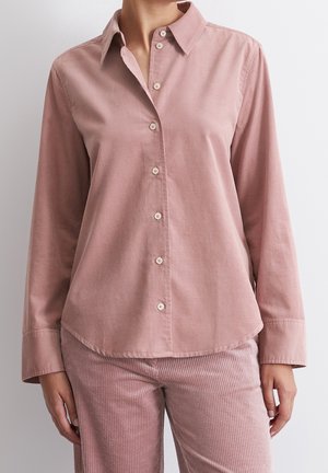 Langarmige stofroze blouse met knopen, gecombineerd met bijpassende roze ribfluwelen broek bij een persoon tegen een effen lichte achtergrond.