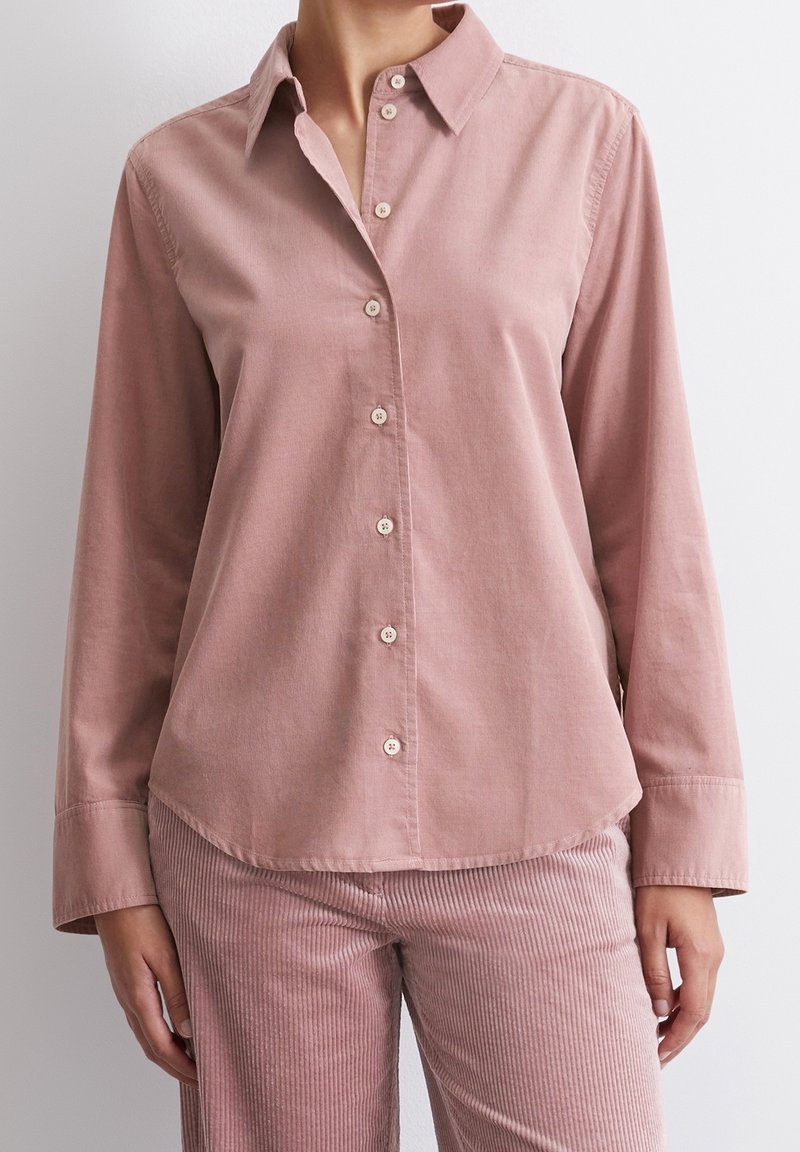 Chemise rose poussiéreuse à manches longues boutonnée, associée à un pantalon en velours côtelé rose assorti, portée par une personne sur un fond clair uni.