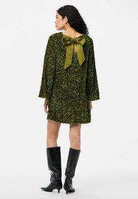 Zaļa spīdīga kleita ar garām piedurknēm, atvērtu muguru un lielu olīvzaļas krāsas loku akcentu. Modelis valkā melnas ceļgalu augstuma zābakus ar krokodila faktūru.