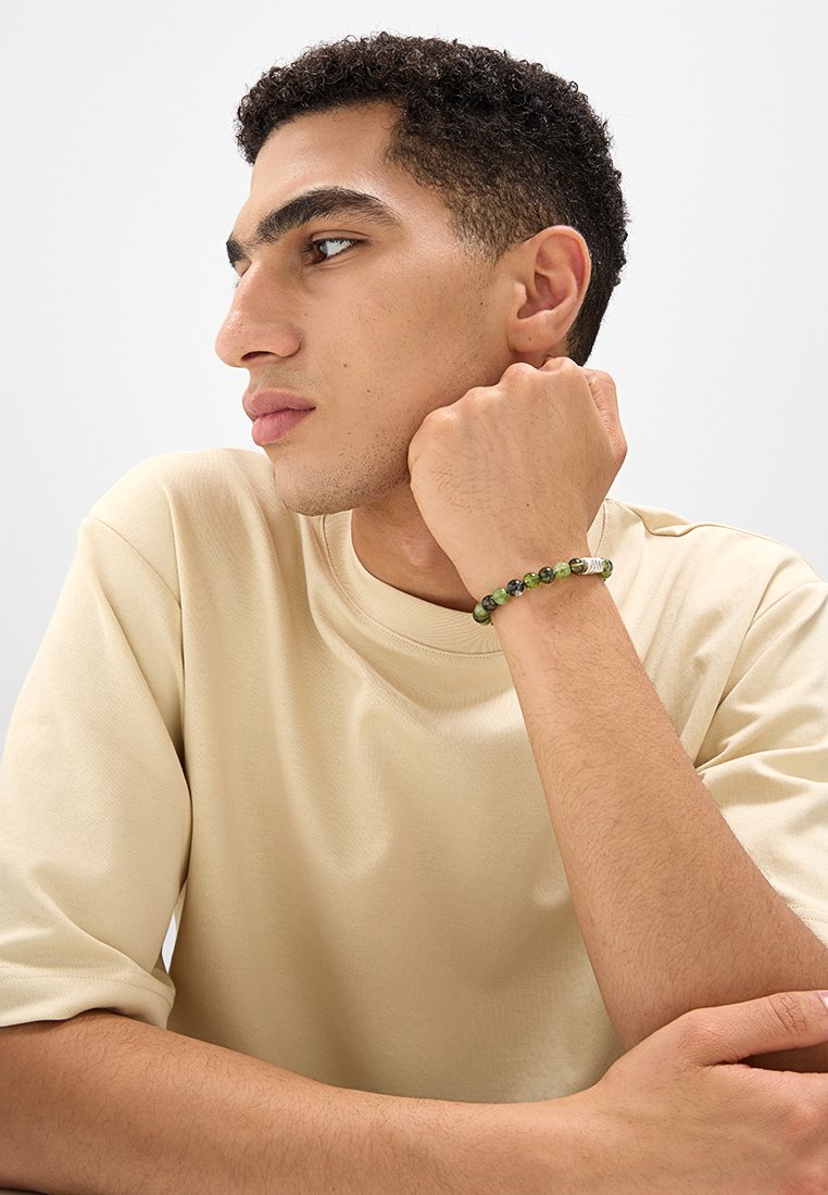 Bracciale di perline verdi con pietre rotonde di diverse sfumature. I dettagli includono un accento metallico. Indossato su una mano appoggiata al volto.