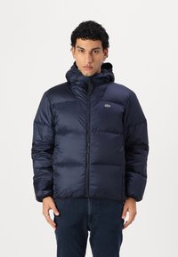Námornícka pufrová bunda s kapucňou, so zipsovým zapínaním a prešívaným dizajnom. Logo Lacoste na ľavej hrudi, hladký povrch a jemná textúra.