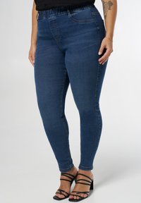 Femme portant un jean skinny bleu foncé et des sandales à talons noires à lanières, se tenant devant un fond clair et uni.