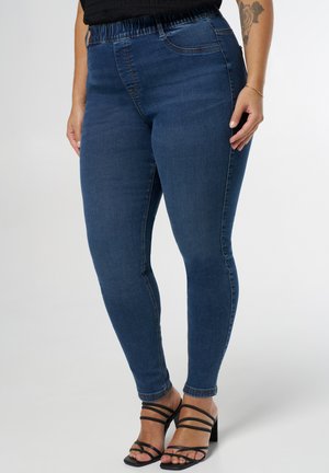 Vrouw in donkerblauwe skinny jeans en zwarte sandalen met hakken, staande tegen een lichte effen achtergrond.