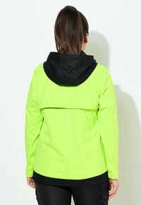 Kirkkaan neonvihreä takki, jossa on musta huppu. Ominaisuuksiin kuuluvat pitkät hihat, rento istuvuus ja kaareva helma. Mukana logo-ominaisuus lähellä keskiselkää.