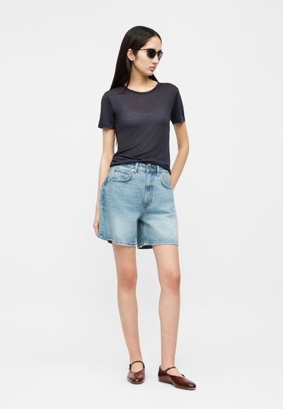 Vrouw draagt zwarte zonnebril, doorschijnend zwart T-shirt, lichtblauwe spijkershorts en bruine Mary Jane schoenen, staand met één hand in de zak.