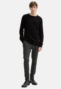 Schwarzer Strickpullover mit rundem Halsausschnitt, kombiniert mit grauen karierten Hosen und schwarzen Schnürschuhen. Glatte Stofftextur sichtbar.