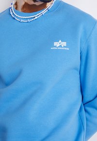 Hombre vestido con un sudadero azul de Alpha Industries y collares de cuentas azules y blancas en capas, recortado en el cuello y el torso.