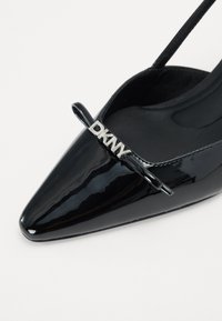 Črne patent usnjene slingback čevlje s špičastim nosom, ki vsebujejo srebrni logotip "DKNY" na trakcu čez prednji del. Gladka tekstura.