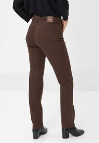 Pantalons marron à jambes droites avec une texture lisse, dotés de poches arrière et d'une étiquette sur la taille. Portés avec des bottines noires.