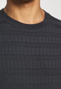 Ciemnogranatowy dzianinowy sweter z kwadratowym, teksturowanym wzorem i prążkowanym dekoltem. Zbliżenie na misterną fakturę tkaniny.