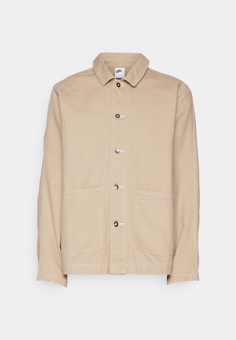 Chaqueta de lona beige con cuello, cierre delantero de botones y dos grandes bolsillos frontales. Presenta un pequeño logo de Nike encima del bolsillo derecho.