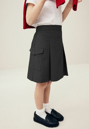 Falda gris con pliegues, un bolsillo lateral y detalle de botón, combinada con mocasines negros y calcetas blancas hasta el tobillo, llevada con un suéter rojo.