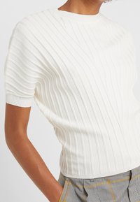 Top corto de punto acanalado blanco con cuello redondo y superficie texturizada. Ajuste ceñido sin características destacadas y sutiles rayas verticales.