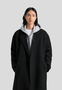 Zwarte wollen jas met een getailleerde pasvorm, voorzien van een gesneden kraag en twee voorkoffers. Gelaagd over een grijze hoodie met rits. Soepele textuur.