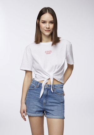 T-shirt in cotone bianco con nodo frontale, con testo rosso "UN DELIRIO ALLA VOLTA GRAZIE." Abbinata a pantaloni corti in denim blu con orli arrotolati.
