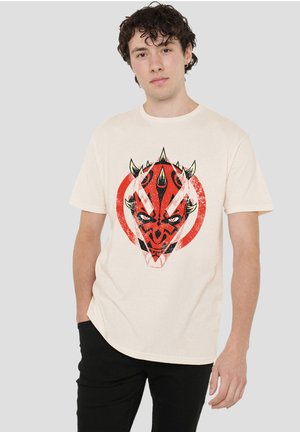 Young man standing, wearing a white t-shirt with a red and black spiked mask graphic and black pants against a plain background.