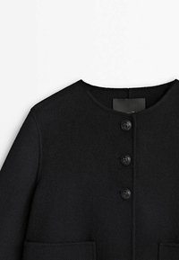 Manteau en laine noire avec encolure ronde, trois boutons sur le devant et deux poches plaquées à l'avant sur un fond clair.