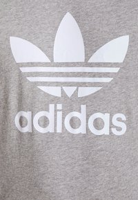 Šedé bavlněné tričko s velkým bílým logem Adidas trefoil a názvem značky v tučném bezpatkovém písmu.