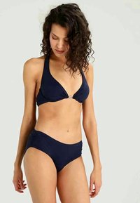 Kvinna med lockigt hår som bär en marinblå halterneck-bikini, tittar ner med ett neutralt uttryck mot en enkel ljus bakgrund.