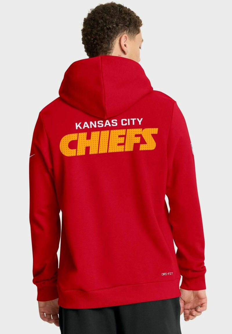 Mann in rotem Hoodie mit dem Text "Kansas City Chiefs" in Gelb und Weiß auf dem Rücken, der vor einem schlichten hellen Hintergrund steht.