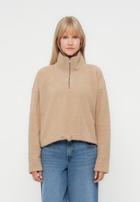 Beiger Fleece-Pullover mit halbem Reißverschlusskragen, überschnittenen Schultern und verstellbarem Kordelzugsaum. Weicher Stoff, lockere Passform.