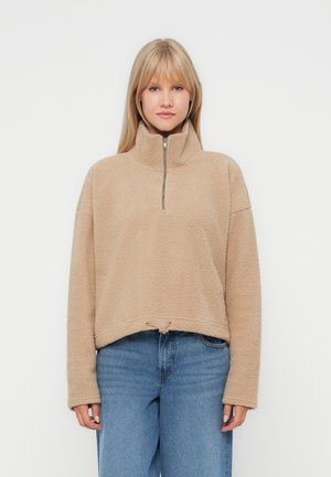 Beige fleece pullover med halvlynlåskrave, dropped shoulders og justerbar snor i kanten. Blød tekstur, afslappet pasform.