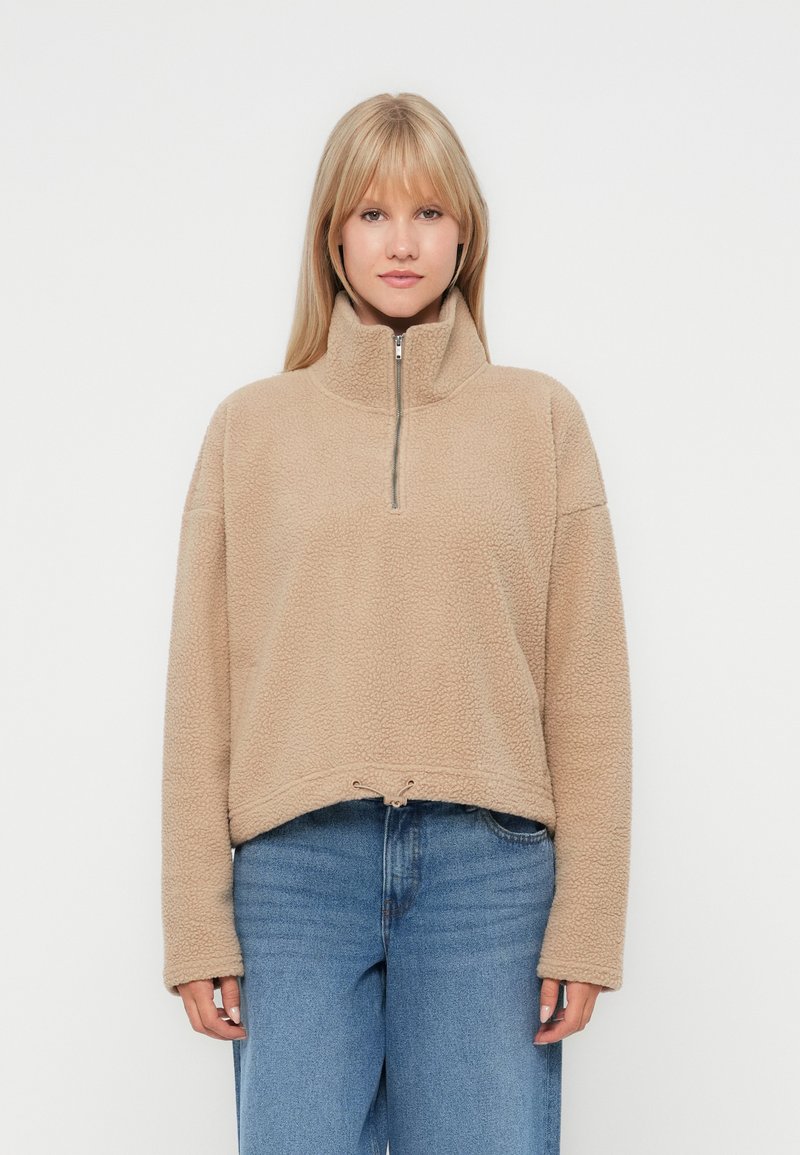 Beiger Fleece-Pullover mit halbem Reißverschlusskragen, überschnittenen Schultern und verstellbarem Kordelzugsaum. Weicher Stoff, lockere Passform.