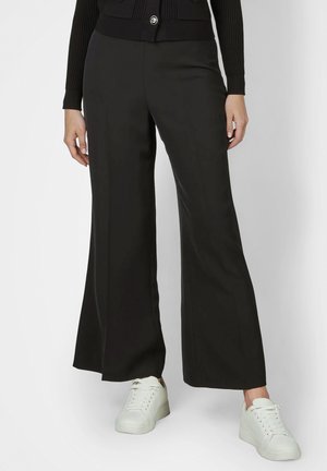 Schwarze Hose mit weitem Bein, kombiniert mit weißen Plateausneakers, gezeigt an einer stehenden Person mit locker hängenden Händen.