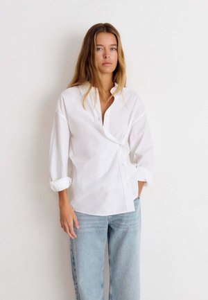 Mujer con cabello largo castaño claro que lleva una camisa blanca holgada abotonada y jeans azul claro, de pie frente a una pared blanca lisa.