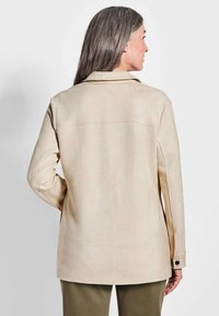 Beige faux suede jas met lange mouwen, een kraag en een rechte snit. Kenmerkt zich door een rugnaad en zwarte knoopaccenten op de manchetten.