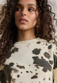 Lichtbeige sweatshirt met een donker spetterpatroon, geribbelde ronde hals en een zachte textuur. Krullend haar en natuurlijke make-up zichtbaar.