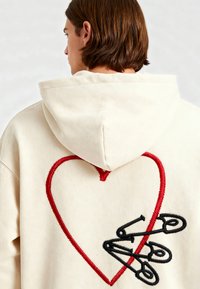 Felpa cremaliera con un grande cuore rosso ricamato e un design di spilla da balia nera. Presenta un cappuccio con cordini e un tessuto texturizzato.