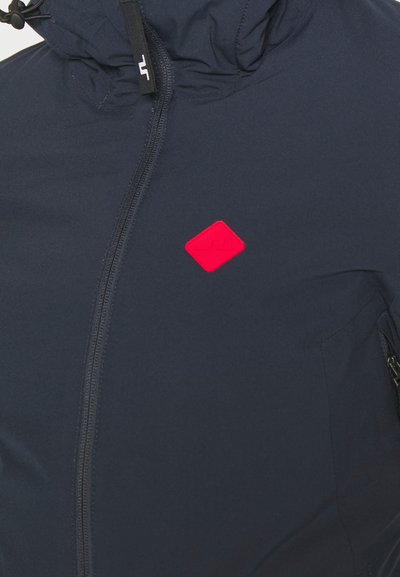 Casaco azul marinho com fecho e um logótipo vermelho em forma de diamante no peito, e capuz ajustável. Apresenta uma textura suave e um design minimalista.